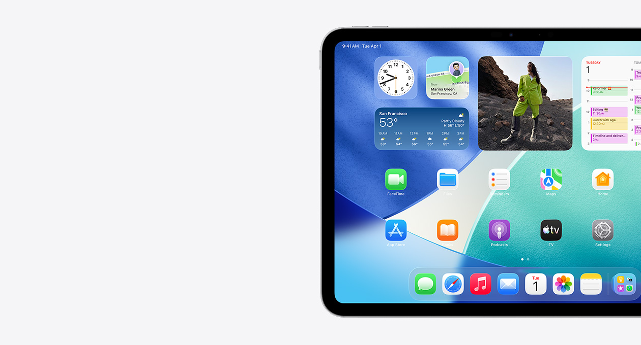 iPad, orientasi lanskap, sisi kiri layar saja, Layar Utama dengan susunan widget yang meliputi jam tembus cahaya yang menampilkan efek Liquid Glass, aplikasi Lacak, widget Cuaca, dua baris aplikasi yang lebih kecil, dan dock