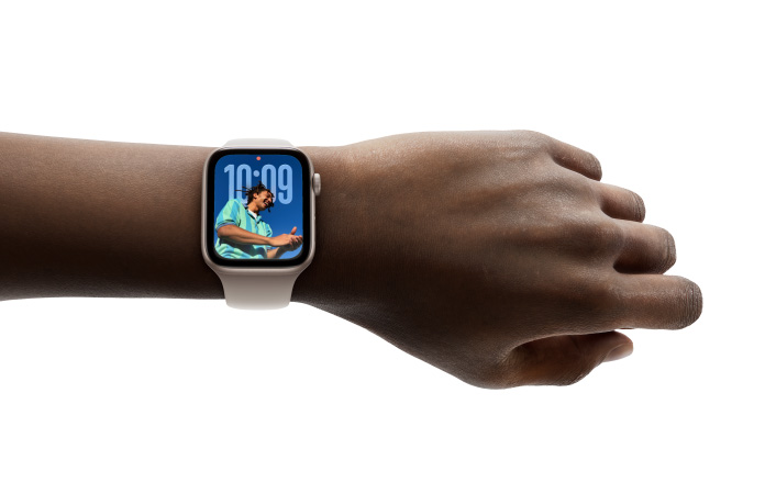 Foto close-up Apple Watch berwarna Perak dengan tali jam karet di pergelangan tangan seseorang, menampilkan aplikasi Waktu dalam desain Liquid Glass baru