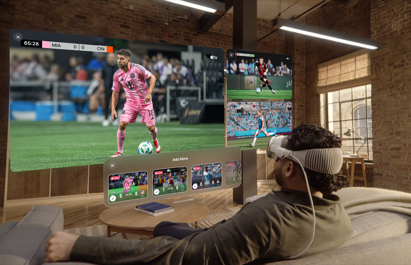 A person wearing Apple Vision Pro is sitting on a couch in a lounge room, displayed in front of them is a large video screen alongside two smaller screens, each showing a different sporting event. Below the large screen is a selection window that reads: Add More