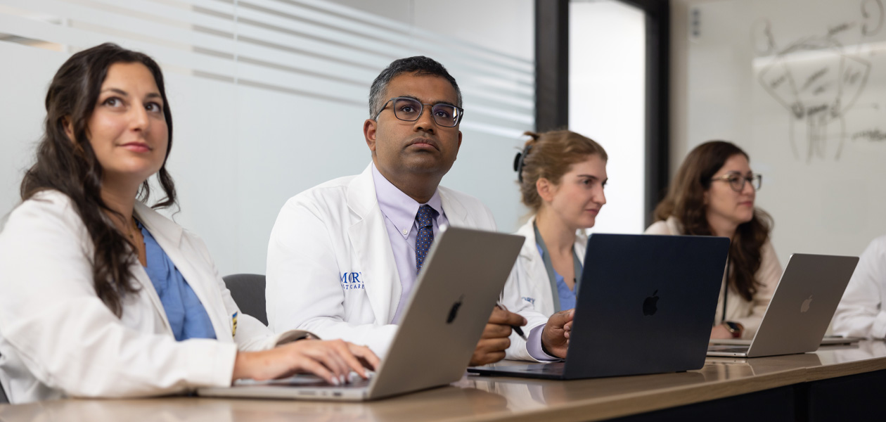 Cuatro personas con batas de médico se sientan en fila. Todas trabajan con un MacBook.
