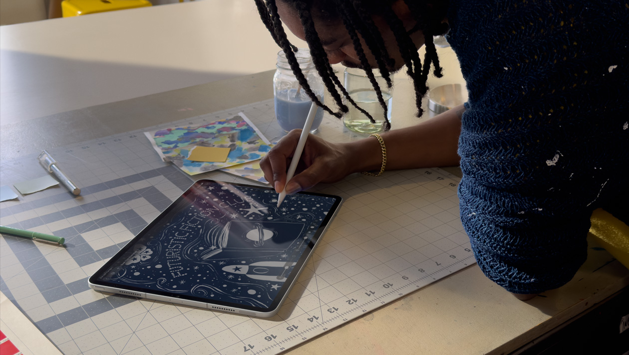 Person mit Braids und blauem Pullover zeichnet auf einem iPad Pro mit Apple Pencil. Auf dem Display ist eine Illustration eines Nachthimmels mit einem Buchcover mit Planeten darauf zu sehen, dazu ein Raumschiff, ein Schmetterling und der Schriftzug „Futuristic Fiction Club“ in der Mitte.